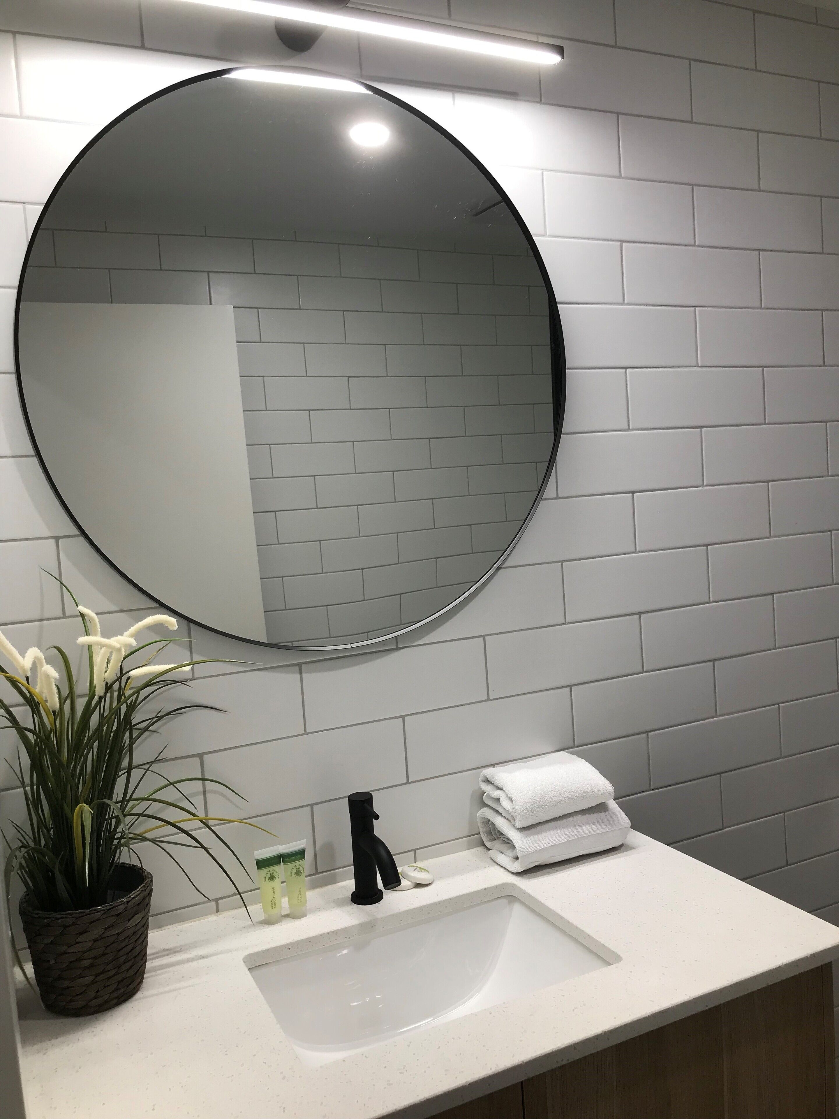 A bathroom with a round mirror, white tiled walls, a sink, a potted plant, and a stack of white towels.