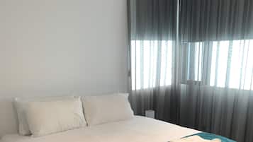 A neatly made bed with white linens and a teal accent, a bedside table with a lamp, and a window with sheer curtains allowing natural light.