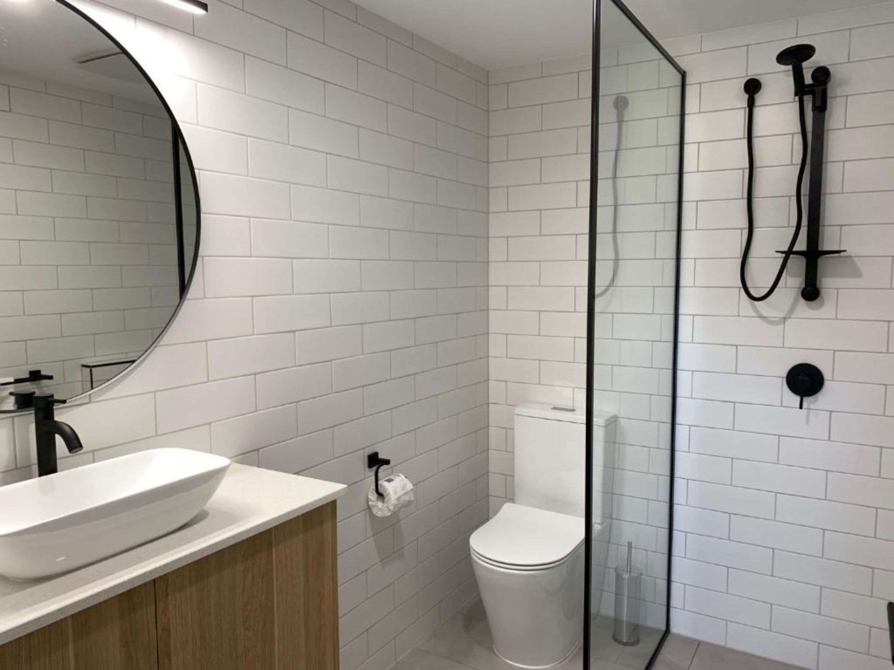 Un baño moderno con pared de azulejos blancos, un espejo redondo, un lavabo blanco y cabina de ducha con mampara de cristal.