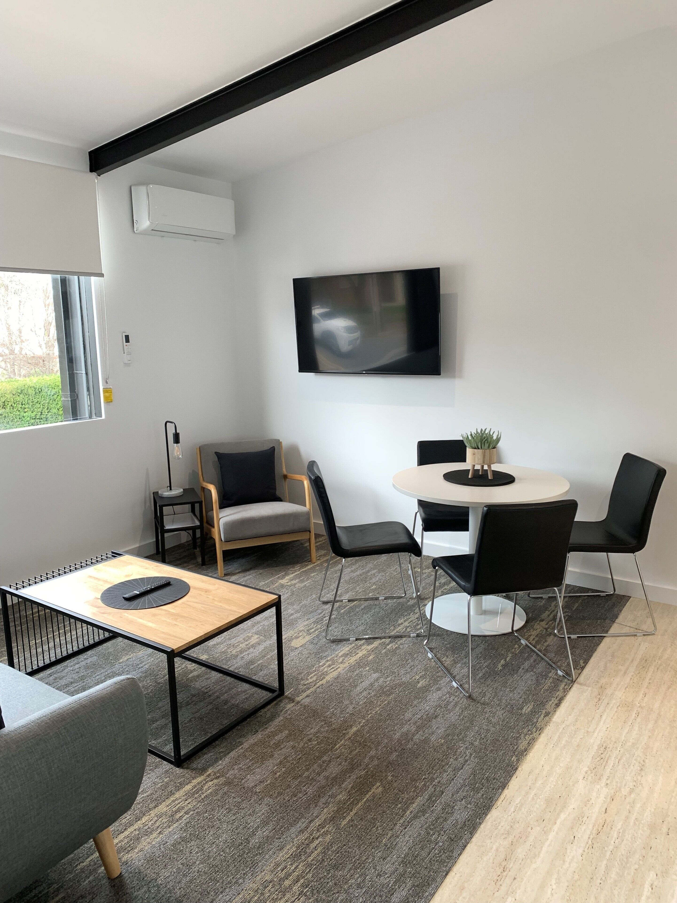 A modern living room with a flat-screen TV mounted on the wall, a round dining table with four chairs, a sofa, and a coffee table.