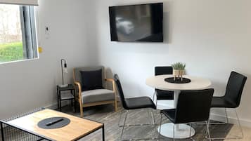A modern living room with a flat-screen TV mounted on the wall, a round dining table with four chairs, a sofa, and a coffee table.