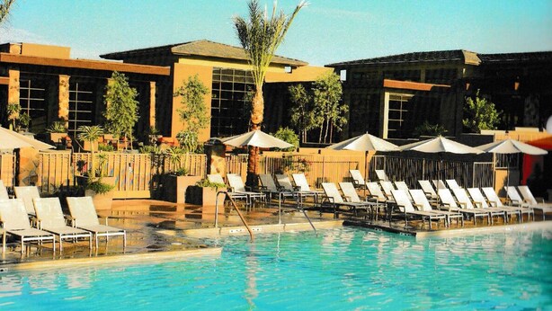 Zona de piscina con sillas de descanso y sombrillas, un edificio con grandes ventanales y palmeras.