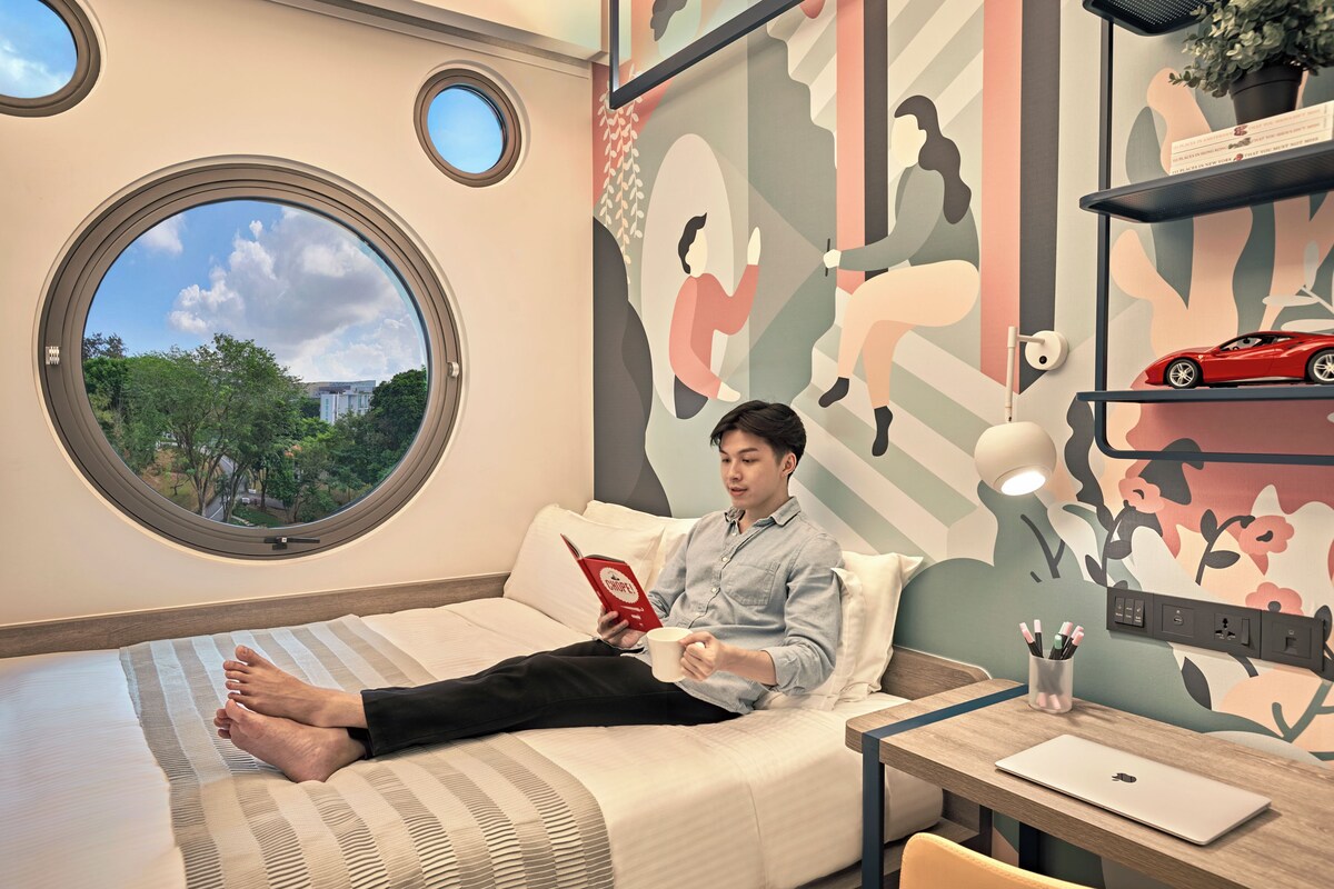 Ein Schlafzimmer mit einem Bett, einem Rundfenster, einem Schreibtisch mit einem Laptop und einer Wand mit abstrakter Kunst.