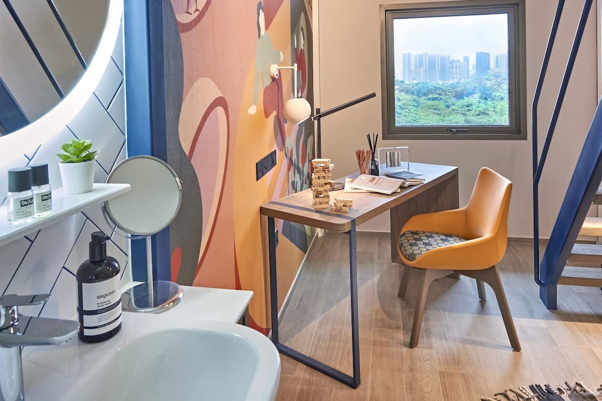 Ein modernes Badezimmer mit großem Spiegel, Waschbecken und einem Sessel. Fenster bieten Blick auf die Stadt, und an der Wand befindet sich ein farbenfrohes Wandgemälde.