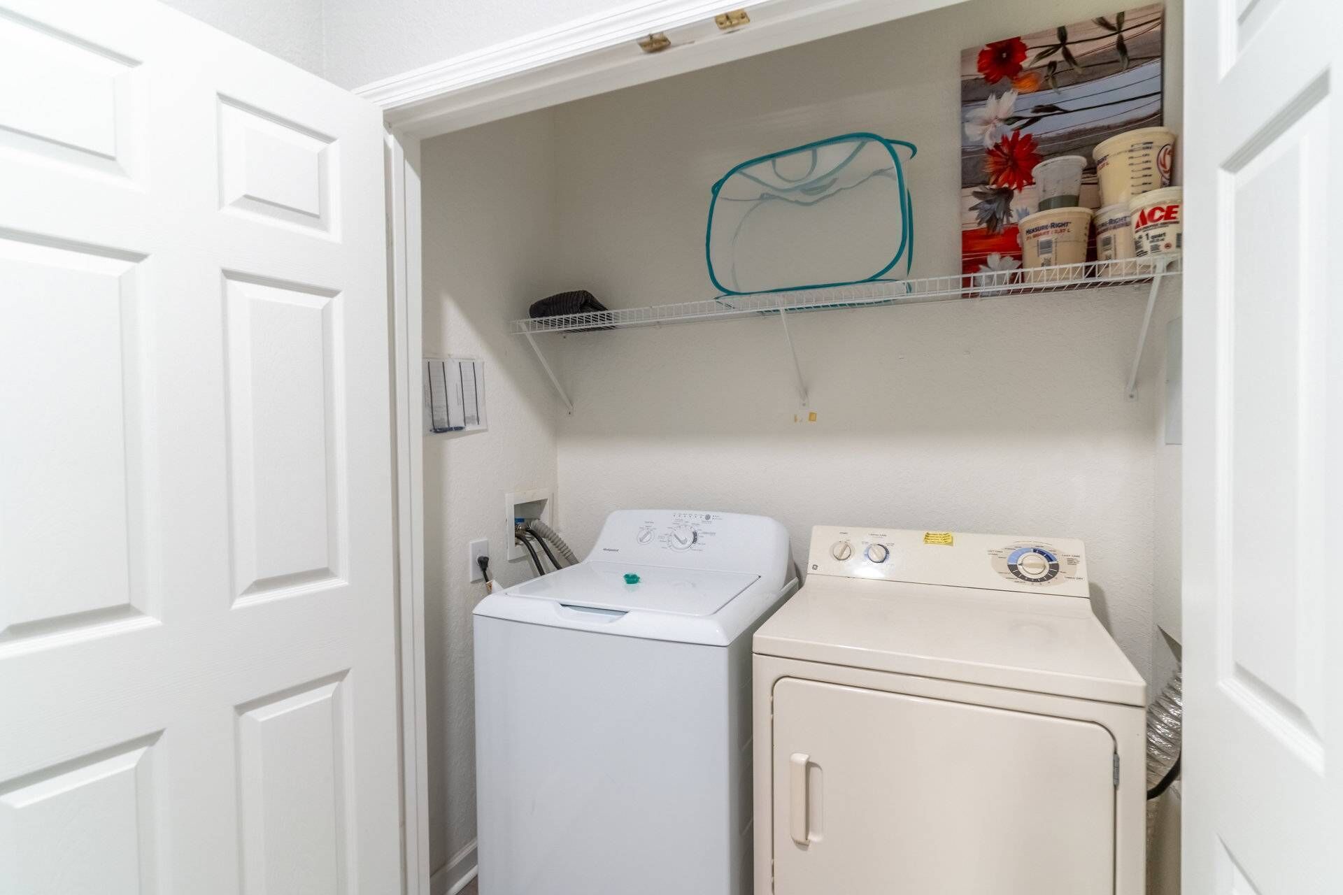 A laundry room with a front-loading washer and a top-loading dryer, a shelf with cleaning supplies, and a door leading to another room.