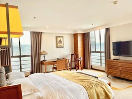 Una habitación de hotel con cama, escritorio, silla, televisor y ventanas amplias.