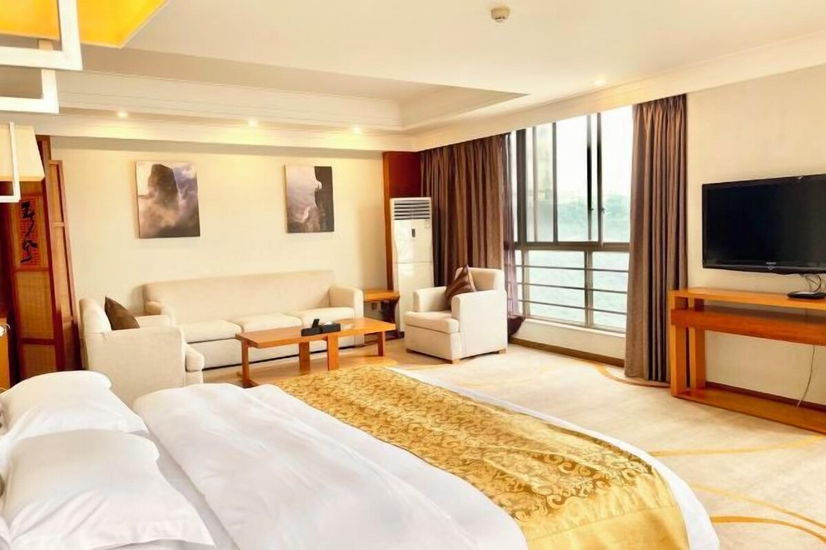 A hotel room with a large bed, a sofa, a coffee table, a TV, and a large window.