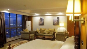 Quarto de hotel com uma cama grande, duas poltronas, um sofá, uma escrivaninha e uma mesa de cabeceira. Há uma janela grande com vista para a cidade à noite.