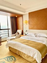 A hotel room with a large bed, a chair, a nightstand, and a view of the city through a large window.