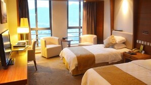 Quarto de hotel com duas camas, uma escrivaninha com candeeiro, uma cadeira e vista para o mar.