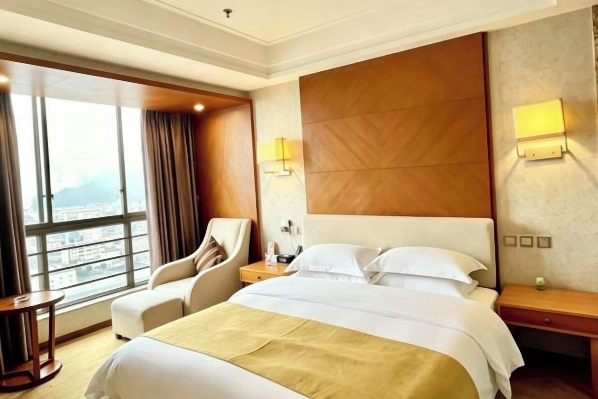 A hotel room with a large bed, a chair, a nightstand, and a view of the city through large windows.