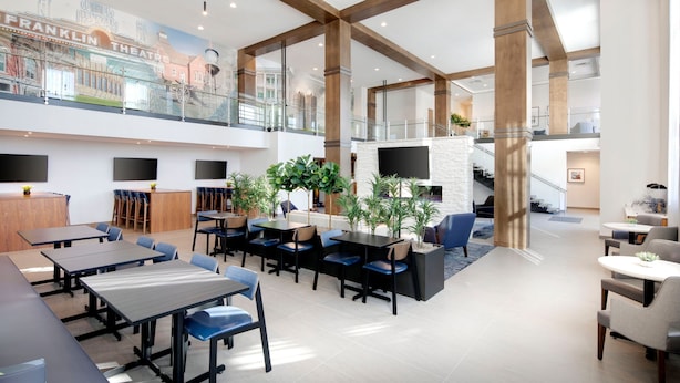 Un hall d’entrée spacieux avec un plafond haut, des poutres en bois, un espace de détente central avec des tables et des chaises, un escalier et une fresque murale.