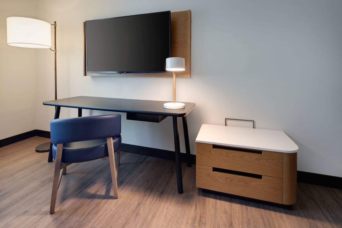 Una habitación moderna con escritorio, silla, televisión, lámpara y una mesita de noche.