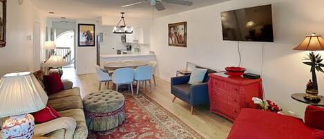 A living room with a sofa, ottoman, and a red couch. There is a dining area with chairs and a table. A TV is mounted on the wall.