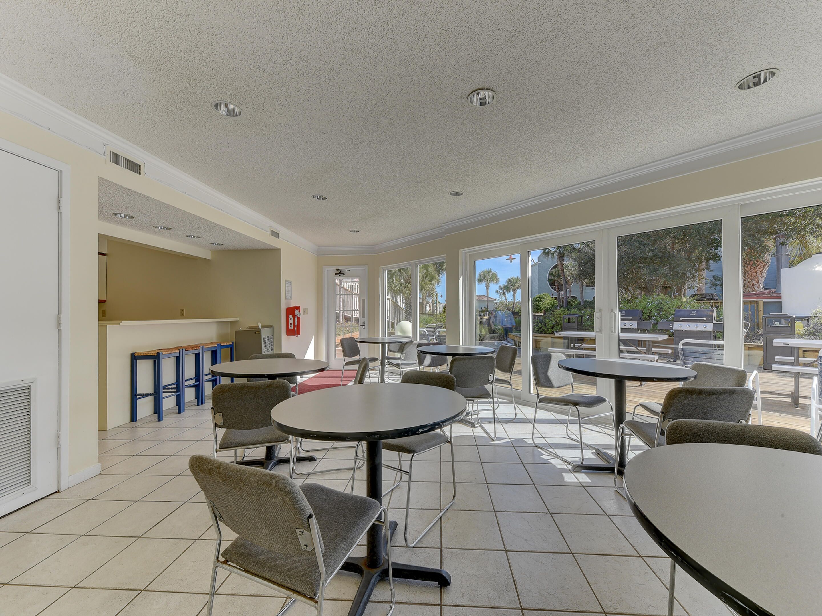 A spacious indoor patio area with multiple round tables and chairs, large windows offering views of the outdoors, and a small kitchenette area.