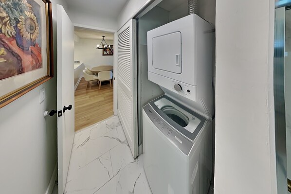 A laundry room with a front-loading washer and dryer, a door with a handle, and a hallway leading to a dining area with a table and chairs.