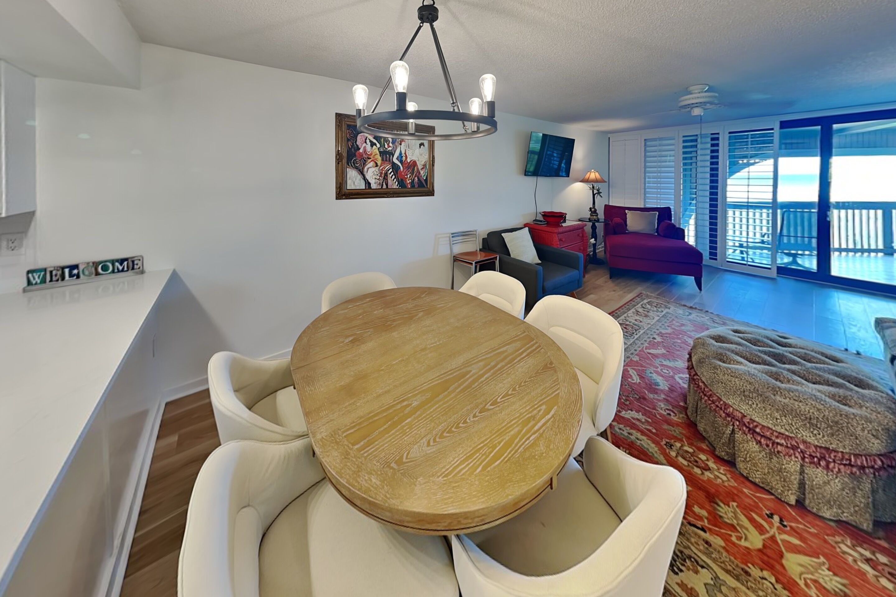 A modern living room with a round wooden dining table, white cushioned chairs, a red sofa, a red ottoman, a TV mounted on the wall, and a balcony with large windows.