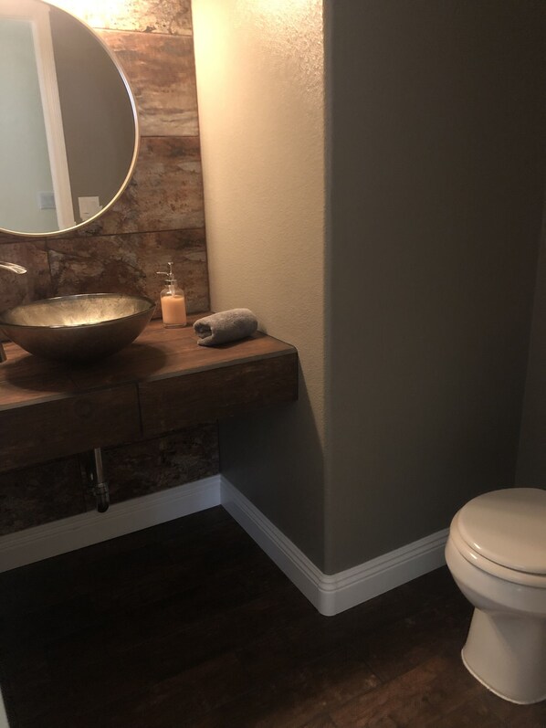 Un bagno con un mobile lavabo in legno, uno specchio rotondo, un lavandino, un asciugamano, un WC e un dispenser di sapone a parete.