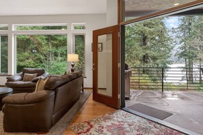 A living room with a brown leather sofa, a small table, and a lamp. There is a large window with a view of trees and a balcony with a railing. - Beautiful Lake Area Home, Gas Grill - Close to Downtown Florence & Dunes (Florence)