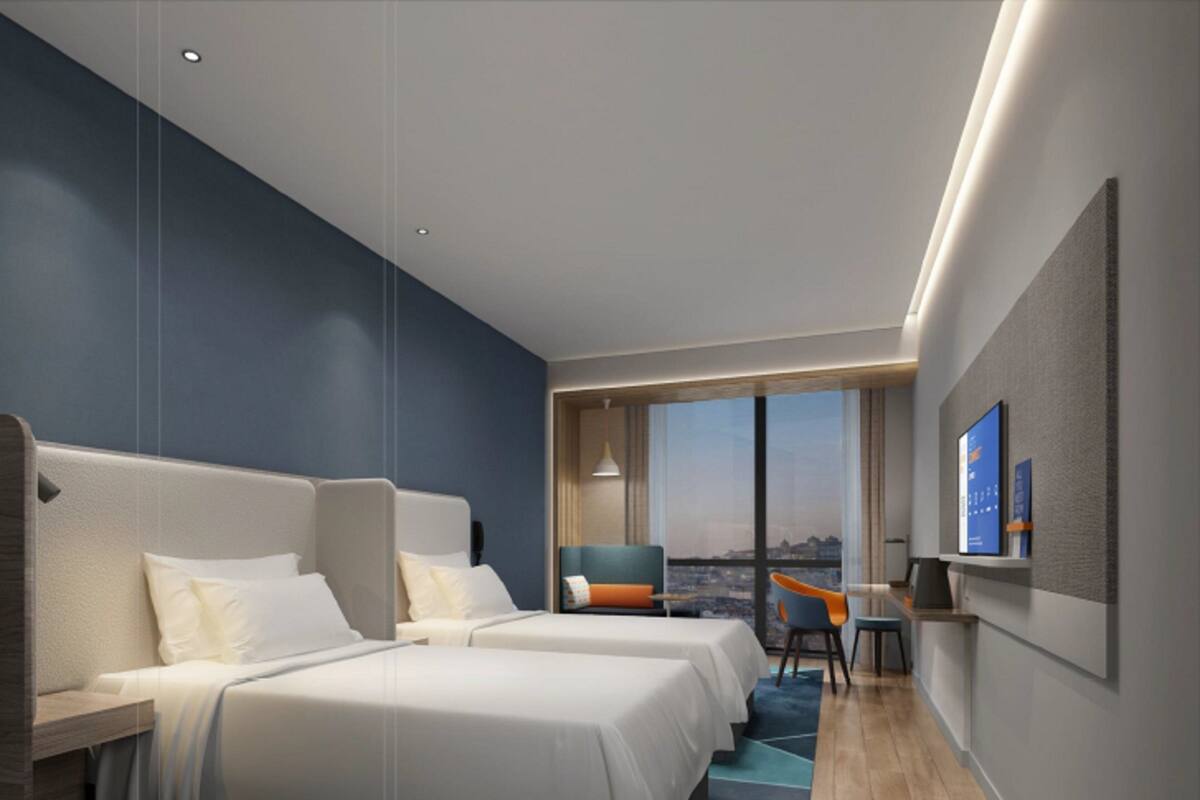 A modern hotel room with a large bed, a desk, and a chair. There is a TV on the wall and a window with a city view.