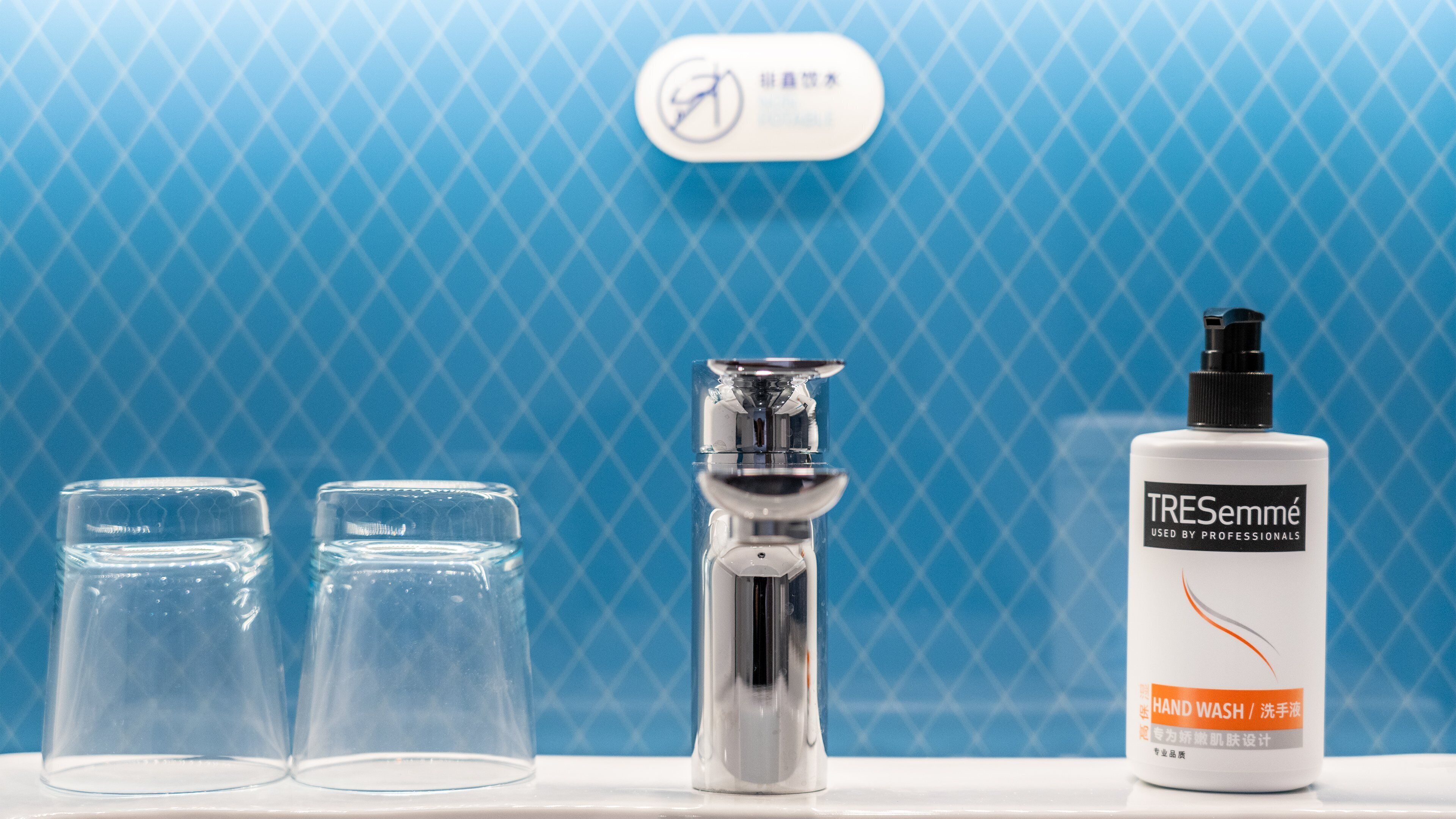 A bathroom with a blue patterned wall, a hand wash bottle, and two clear glasses.