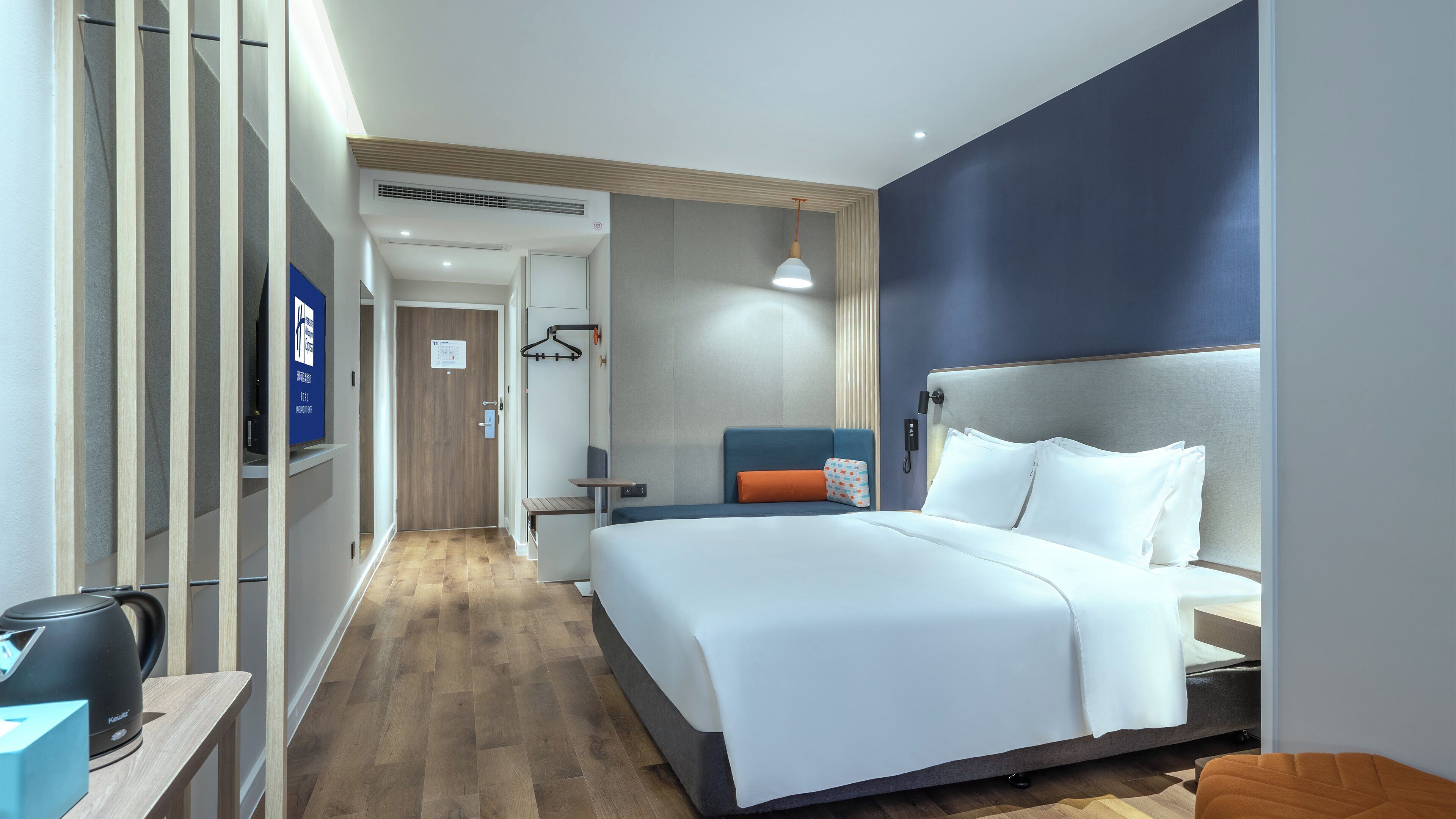 A hotel room with a bed, bedside table, and a seating area with a chair and cushions.