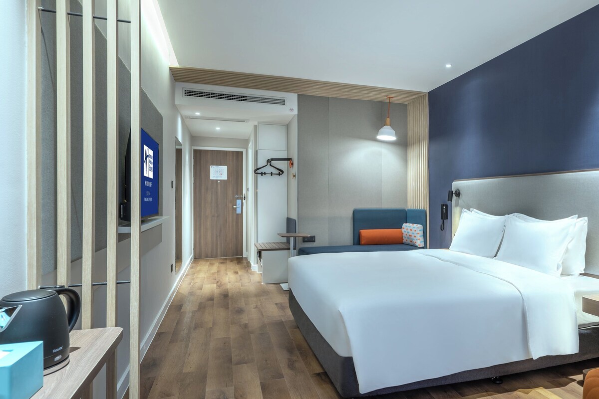 A hotel room with a large bed, a desk with a kettle, and a seating area with a blue cushion.