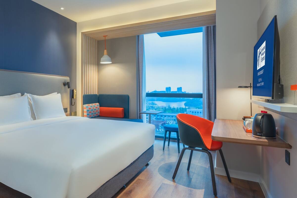 A hotel room with a large bed, a desk with a chair, a coffee maker, and a view of the city through a large window.