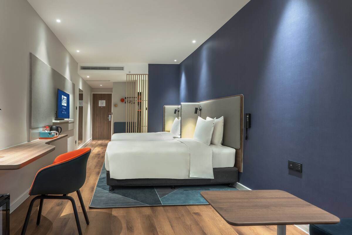 A modern hotel room with a large bed, a desk with a chair, a TV, and a minibar.