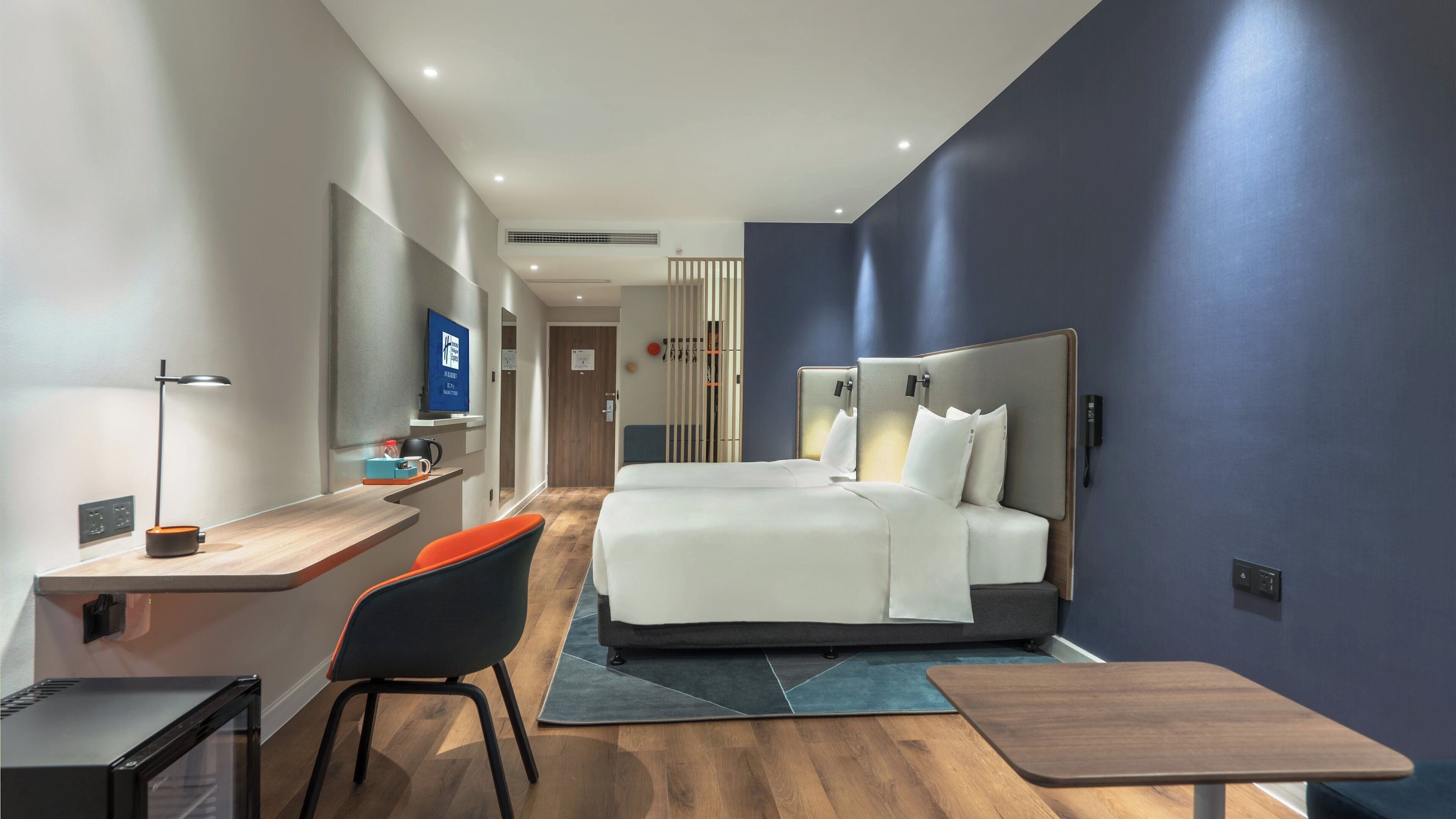 A modern hotel room with a large bed, a desk with a chair, a TV, and a minibar.