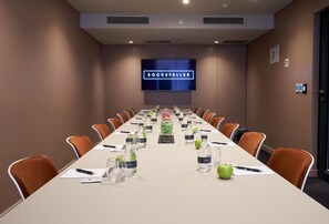 A conference room with a long table, orange chairs, water bottles, and a television screen displaying "ROCKEFELLER". - The Rockefeller Hotel by Newmark (Cape Town)