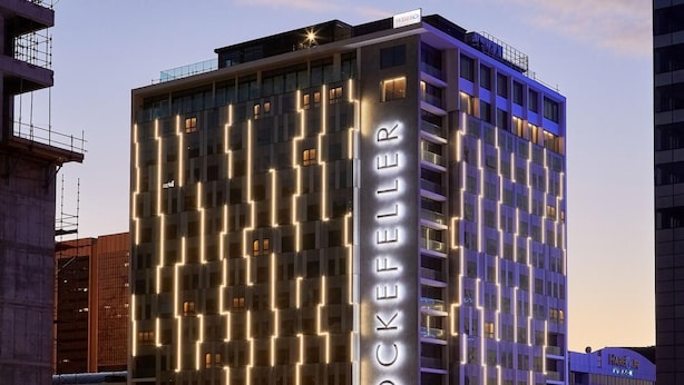 A high-rise hotel with a modern facade featuring illuminated vertical lines and the name "The Rockefeller" prominently displayed. - The Rockefeller Hotel by Newmark (Cape Town)
