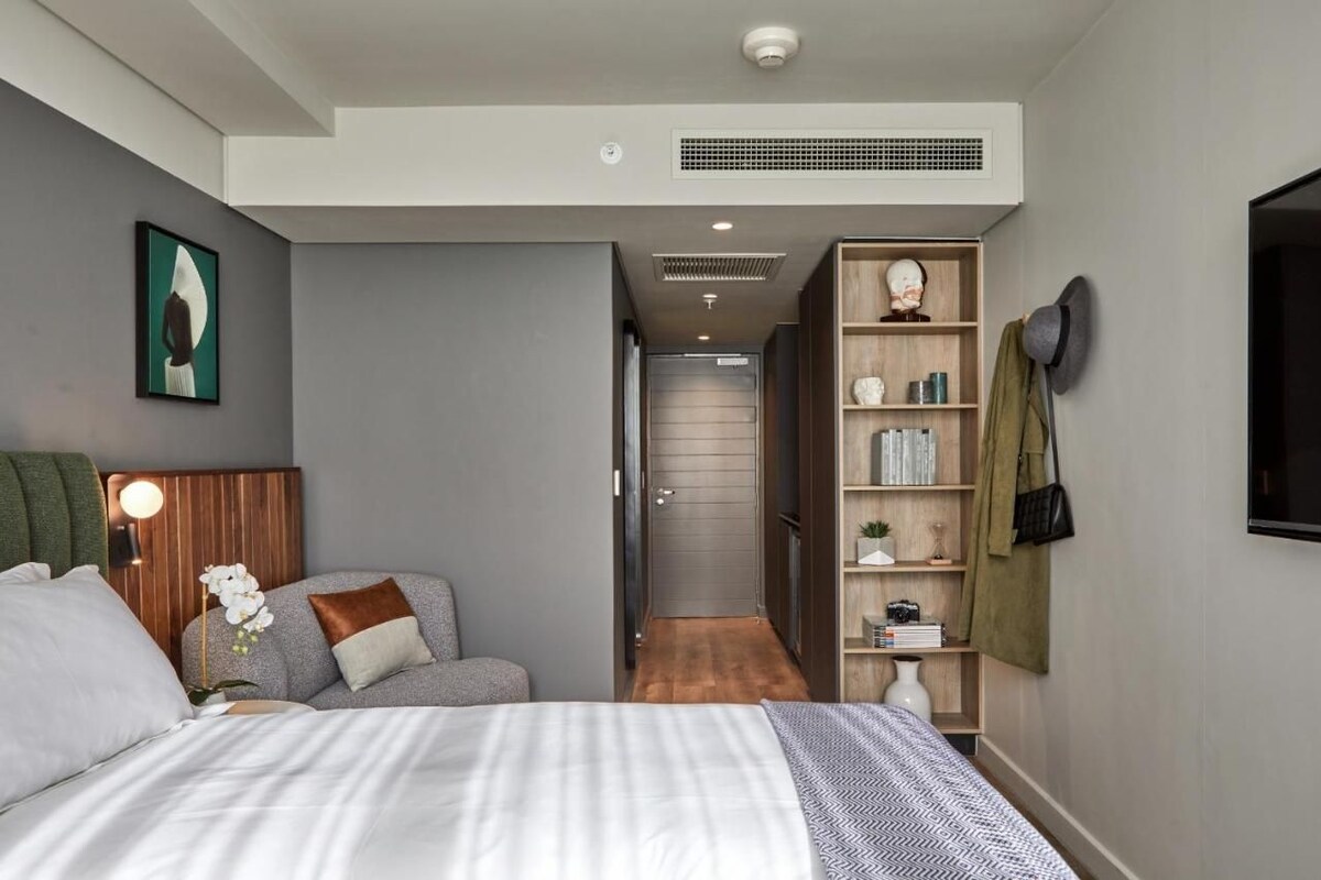 A modern hotel room with a large bed, a seating area with a sofa, a bookshelf, and a wardrobe.