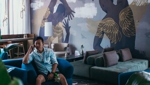 A modern living room with a person sitting on a blue sofa, a large mural on the wall, and a ceiling with a geometric design.