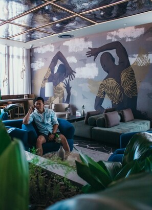 A modern living room with a person sitting on a blue sofa, a large mural on the wall, and a ceiling with a geometric design. - The Rockefeller Hotel by Newmark (Cape Town)