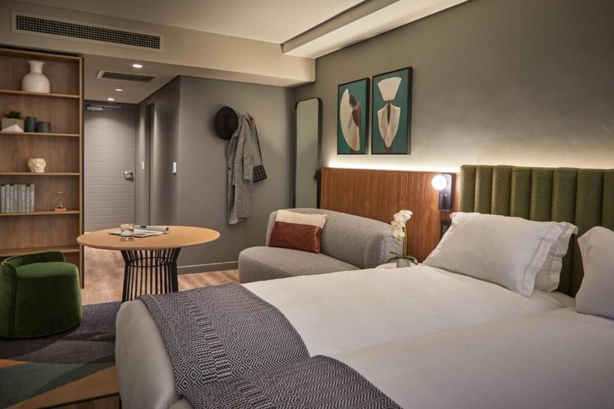 A modern hotel room with a bed, a round table, a bookshelf, and a green armchair.