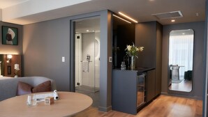 A modern hotel room with a round table, a small bar area, and a shower room visible through an open door.