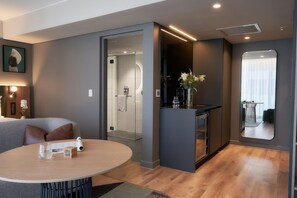 A modern hotel room with a round table, a small bar area, and a shower room visible through an open door. - The Rockefeller Hotel by Newmark (Cape Town)