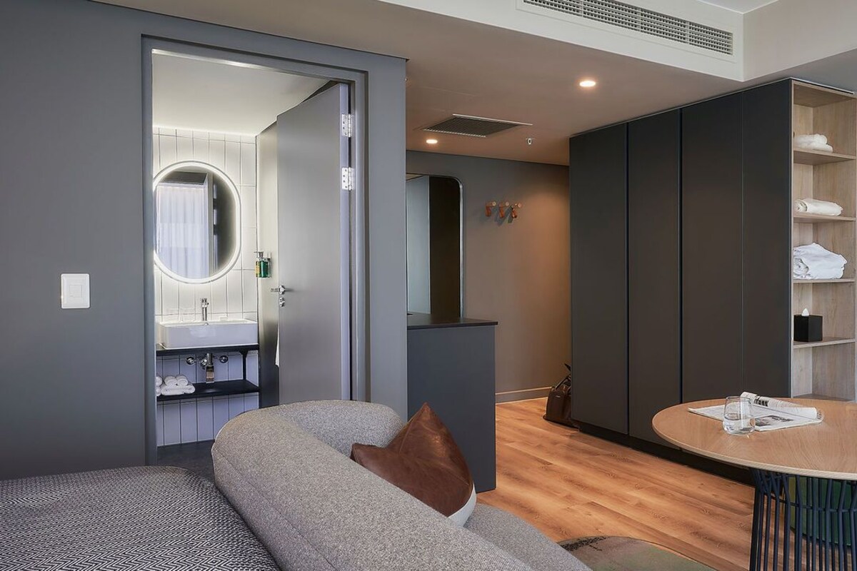 A modern living room with a grey sofa, a round wooden table, and a built-in kitchen area with a sink and mirror.