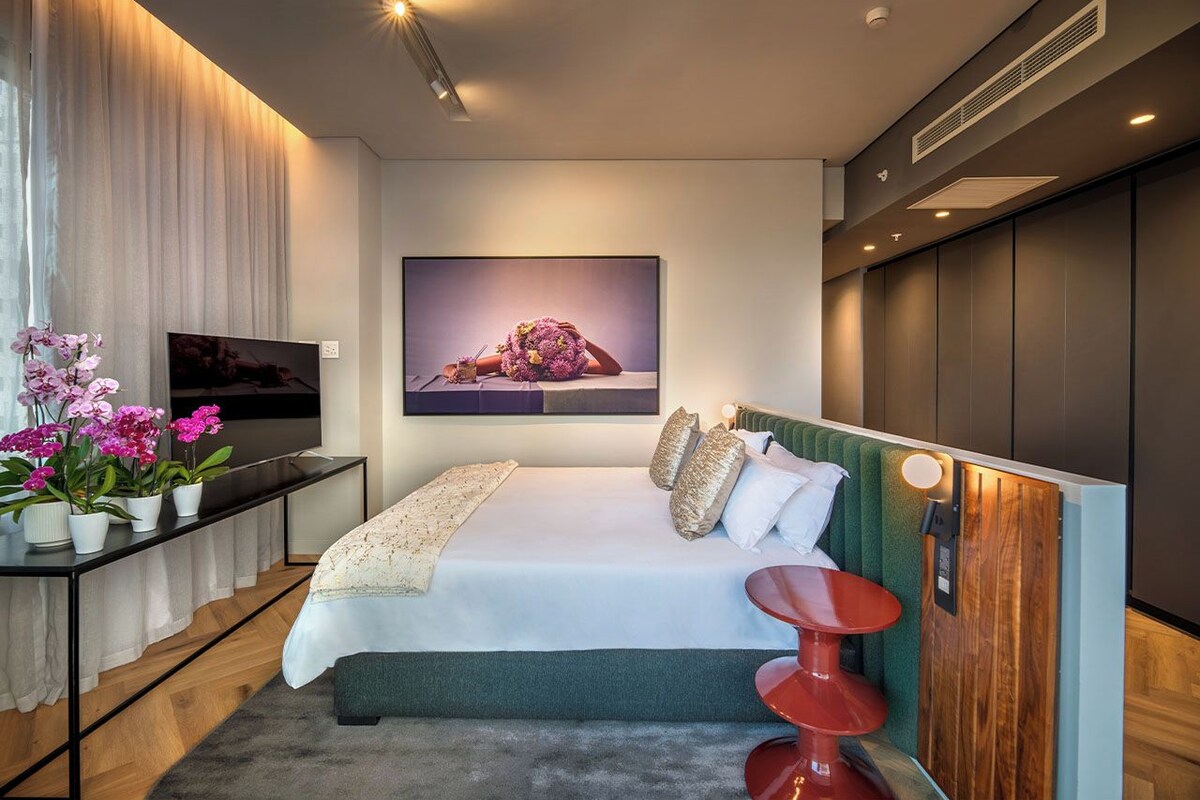 A modern bedroom with a bed, a TV, a red chair, and a wall-mounted artwork.