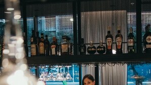 A bar area with a bartender, shelves of liquor bottles, and a view of the cityscape through the window. - The Rockefeller Hotel by Newmark (Cape Town)