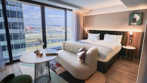 A modern hotel room with a large bed, a sofa, a small round table, and a view of the cityscape through large windows. - The Rockefeller Hotel by Newmark (Cape Town)