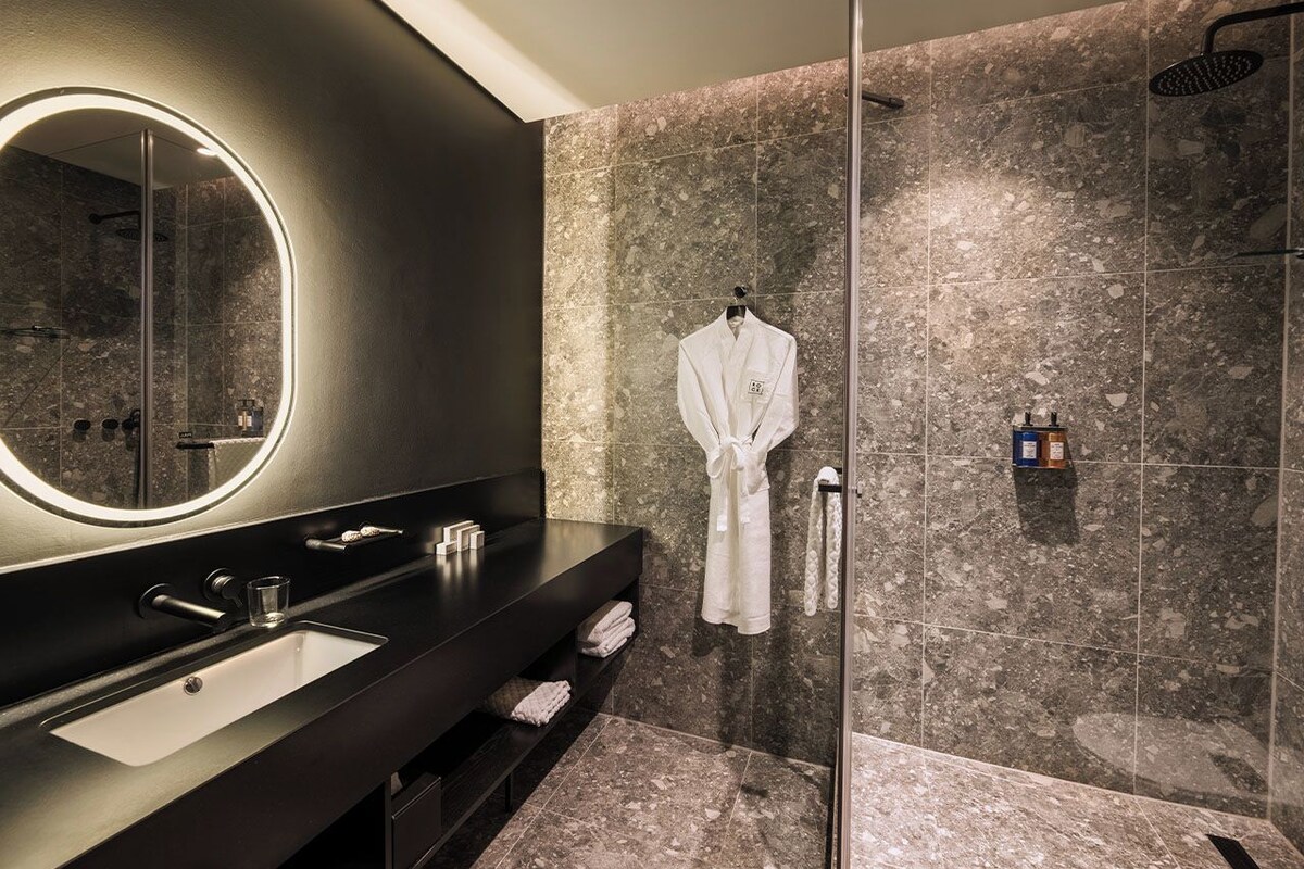 A modern bathroom with a dark vanity, a round mirror, a glass shower enclosure, and a robe hanging on the wall.