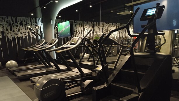A row of stationary bikes and treadmills in a gym with a modern design, including a digital screen and a wall with a unique pattern.