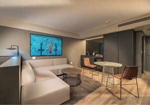 Ein modernes Wohnzimmer mit einer Couch, einem runden Couchtisch und zwei Sessel. Im Hintergrund ist eine Küchenzeile mit einem Esstisch und Stühlen zu sehen.