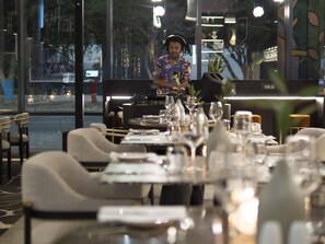 A restaurant interior with tables set for dining, a bartender preparing drinks, and a view of the street outside. - The Rockefeller Hotel by Newmark (Cape Town)