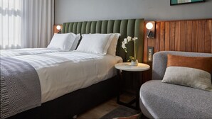 A hotel room with a bed, a chair, a small table with a vase of flowers, and a painting on the wall.