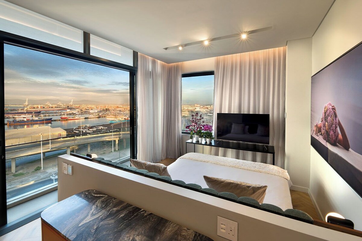 A modern hotel room with a large window offering a city view, a flat-screen TV, and a comfortable bed.