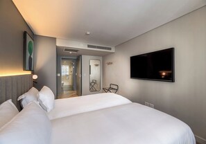 A hotel room with a large bed, a flat-screen TV mounted on the wall, a door leading to a bathroom, and a small table with a chair. - The Rockefeller Hotel by Newmark (Cape Town)