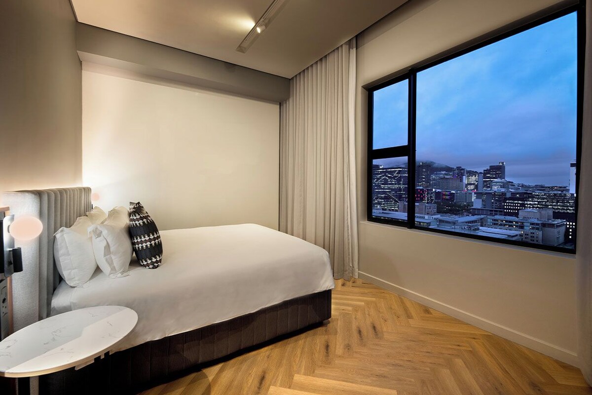A modern hotel room with a large window offering a city view, a bed with white bedding, a round marble-topped table, and a wall-mounted TV.